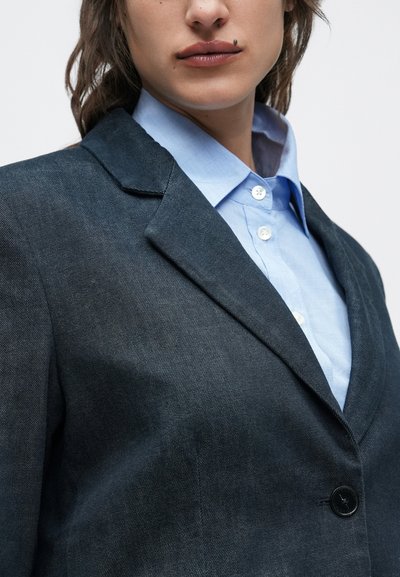 Μαύρο τζιν σακάκι με εντοπισμένα πέτα και ένα κουμπί, φορεμένο πάνω από ένα γαλάζιο πουκάμισο με κουμπιά. Η υφή φαίνεται λεία και προσαρμοσμένη.