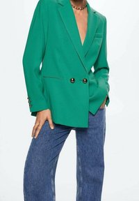 Groene dubbelgeknoopte blazer met grote zwarte knopen, gelapte revers en lange mouwen. Gepaard met blauwe wijde jeans.