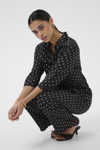 Femme aux cheveux foncés vêtue d'une combinaison noire à motifs, boucles d'oreilles créoles en or, bracelet et sandales noires à talons, accroupie sur un fond blanc.