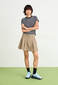 Raitapaita musta-valkoinen, khaki-värinen laskostettu hame, siniset urheilukengät mustilla yksityiskohdilla, seisoo vihreällä pinnalla.