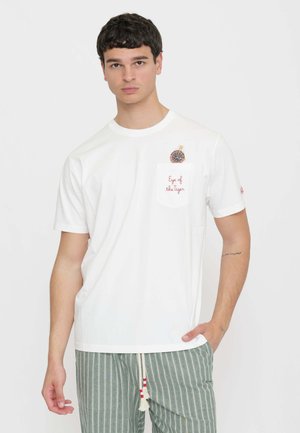 Giovane uomo con capelli corti e scuri indossa una maglietta bianca con una tasca etichettata "Eye of the Tiger" e pantaloni a righe verdi e bianchi.