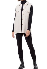 Veste beige sans manches avec quatre poches à boutons, boutons sur le devant et texture douce, portée par-dessus un col roulé noir et des leggings noirs.