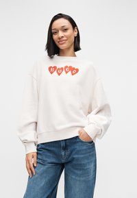 Svetlo bež jopa z rdečimi grafičnimi motivi srca in napisom "HUGO". Ima spuščena ramena in široke rokave, kombinirana z modrimi sproščenimi kavbojkami.