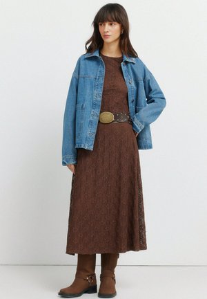 Kobieta w brązowej teksturowanej sukience midi z szerokim paskiem z ćwiekami, niebieskiej kurtce dżinsowej i brązowych botkach do połowy łydki stoi oparta o białą ścianę.