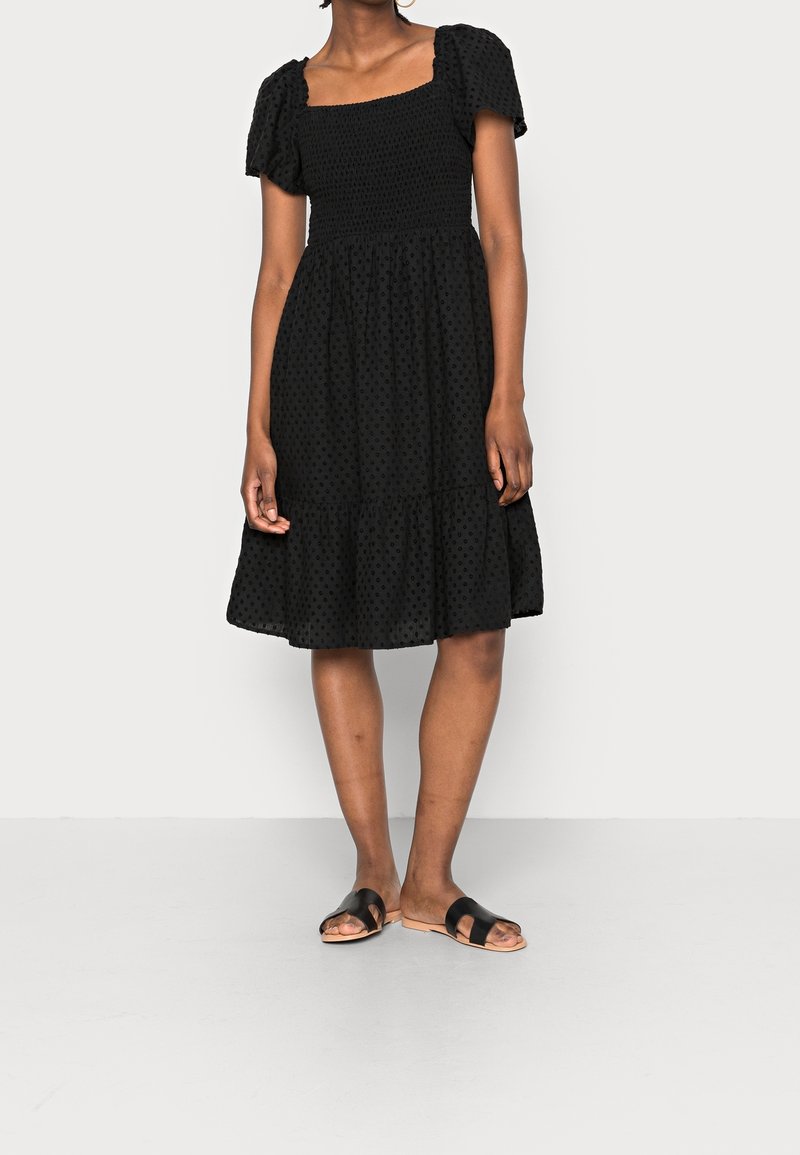 Femme portant une robe noire longueur genou avec des manches bouffantes et un motif à pois texturé, associée à des sandales plates noires sur un fond uni.