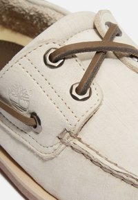 Beige Bootsschuh mit strukturierter Oberfläche, Lederband, Metallösen und kontrastierenden Nähten auf einer braunen Gummisohle.