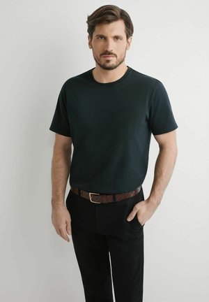 Mężczyzna o krótkich brązowych włosach i brodzie, ubrany w ciemnozieloną koszulkę, czarne spodnie i brązowy pasek, stojący z jedną ręką w kieszeni.