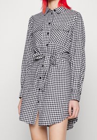 Vestido de camisa a cuadros en blanco y negro con mangas largas, cierre de botones en el frente, dos bolsillos en el pecho y cintura ceñida con cinturón.