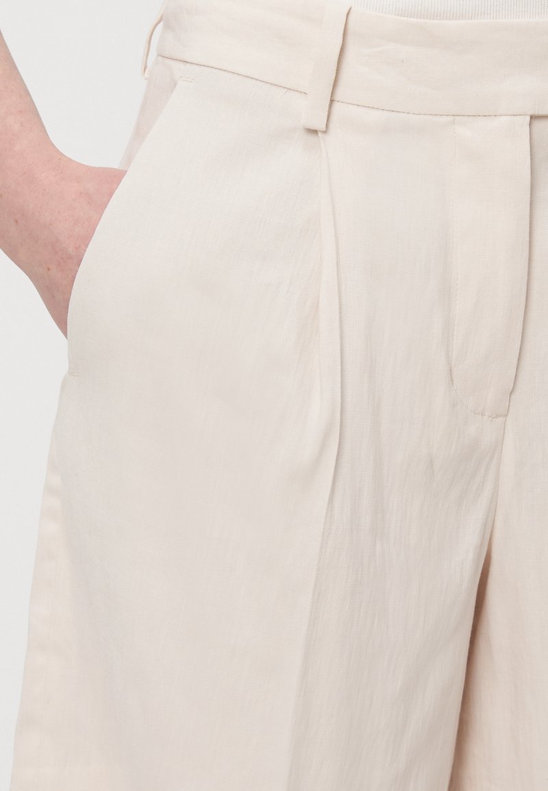 Beige Leinenhose mit einem vorderen Falten, seitlichen Taschen und einem flachen Bund. Die Stoffoberfläche wirkt leicht und leicht zerknittert.