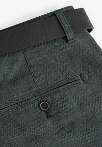 Pantalons en tissu texturé vert foncé avec une ceinture en simili cuir noire, une fermeture à bouton et des détails de poches arrière.
