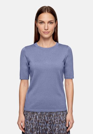 Femme aux cheveux bruns raides portant un pull à manches courtes violet clair et une jupe plissée avec des motifs bleus et gris.