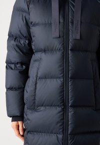Marineblå pufferjakke med kviltet design, frontglidelås og to sidelommer med teksturert stoff og kontrasterende stroppdetaljer.