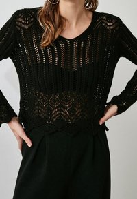 Schwarzer gehäkelter Pullover mit einem lockeren, offenen Strickdesign, langen Ärmeln und gewelltem Saum, kombiniert mit schwarzen hochgeschnittenen Hosen.