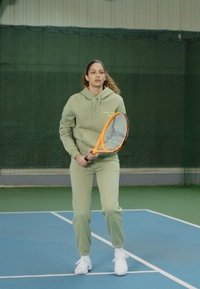 Jogador de ténis com um hoodie verde claro e calças de treino combinando, segurando uma raquete laranja, de pé numa quadra azul com fundo verde.