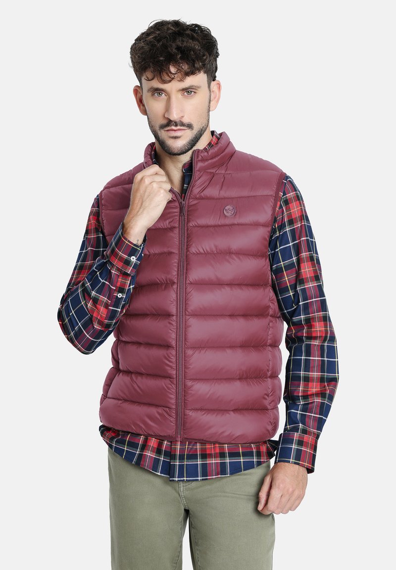 Chaleco de plumas burdeos con textura acolchada vertical, cierre de cremallera y logotipo elevado. Llevado sobre una camisa de cuadros roja y azul. Estilo casual.