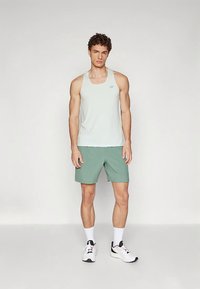Man staat in een wit mouwloos sporttop, groene shorts, witte sokken en witte hardloopschoenen tegen een effen achtergrond.