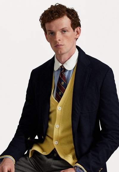 Marineblå blazer over en lyseblå skjorte, gul cardigan og stripete slips. Grå bukser, knappedetaljer på cardiganen og synlig klokke.