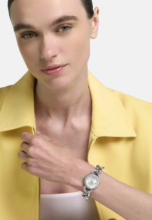 Vrouw met een gele jas en een wit topje, die een zilveren horloge met schakelkettinkje aan haar linker pols toont.