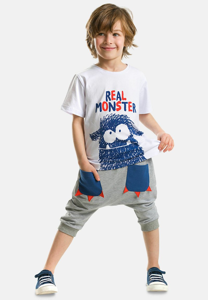 Weißes T-Shirt mit blauer Monstergrafik und rotem Text; graue Jogginghose mit blauen Taschen und roten Dreieckakzenten; blaue Turnschuhe.