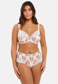Femme portant un ensemble de lingerie en dentelle florale blanche avec des fleurs roses et pêches, se tenant devant un fond clair uniforme.