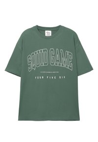 Vihreä puuvillapaita, jossa on teksti "SQUID GAME" paksuilla valkoisilla kirjaimilla ja "FOUR FIVE SIX" alla pienemmällä fontilla. Pyöreä pääntie.