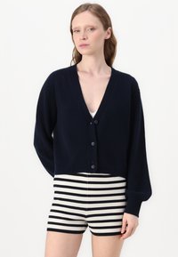Marineblå cropped cardigan med dyb V-hals, knaplukning og ribbede ærmer. Kombineret med ribbede shorts med cremefarve og sorte striber.