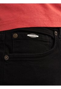 Nahaufnahme einer schwarzen Jeans-Tasche mit Metallknöpfen und einem "PETROL IND"-Etikett, dazu ein sichtbarer Saum eines roten Hemds darüber.