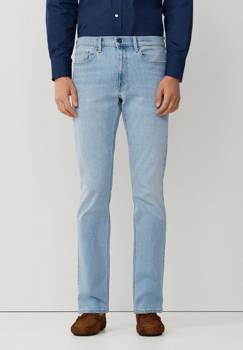 Man met lichtblauwe jeans met rechte pijpen, donkerblauw overhemd met knopen en bruine mocassins, staand tegen een effen achtergrond.