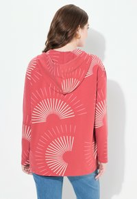 Haut à capuche rouge avec un grand motif en éclat de soleil blanc, manches longues, et un cordon de serrage sur la capuche, associé à un jean bleu. Texture de tissu lisse.