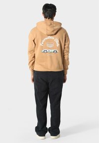 Sudadera de algodón marrón con un gráfico de un coche de carreras vintage en la parte posterior. Combinada con pantalones negros, presenta un corte relajado y capucha ajustable.