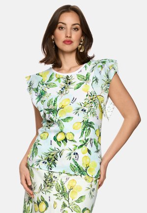 Frau trägt ein weißes Top und einen Rock mit gelbem Zitronen- und grünem Blattmuster, posiert mit einer Hand auf der Hüfte vor einfarbigem Hintergrund.