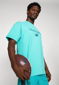 Camiseta turquesa de los Miami Dolphins con logo, de manga corta y cuello redondo; persona sosteniendo un balón de fútbol americano marrón.