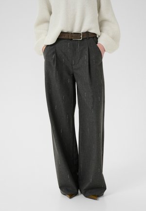 Wide-leg trousers in dark gray fabric featuring vertical pinstripes and pleats, paired with a brown belt and a textured cream sweater.