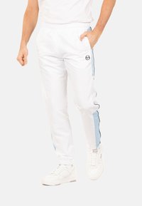 Pantalons de sport blancs avec des rayures latérales bleues et noires, taille élastique et poches latérales, associés à des baskets blanches.