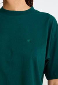 Mørkegrønn T-skjorte laget av bomull, med rund hals og kortermet. Den lille, broderte logoen er plassert på venstre bryst.