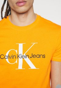 Světlé oranžové tričko vyrobené z bavlny. Na přední straně se nachází velké bílé logo "CK" s textem "Calvin Klein Jeans" v černé barvě pod logem.