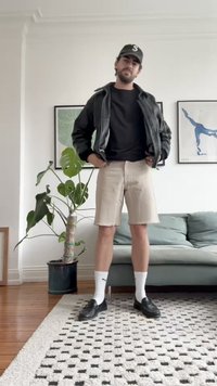 Casaco de pele preto, sweatshirt preta, calções bejes, meias brancas, loafers pretos. De pé numa sala com uma planta verde e um sofá.
