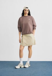 Νεαρή γυναίκα με μπεζ φούστα, βιολετί φούτερ, λευκό καπέλο, λευκές κάλτσες και μπεζ αθλητικά παπούτσια, στέκεται σε μπλε χαλί.