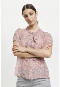 Giovane donna con capelli scuri raccolti, che indossa una blusa a maniche corte rosa con fiori e pantaloni grigi, in piedi con le braccia incrociate su uno sfondo bianco.