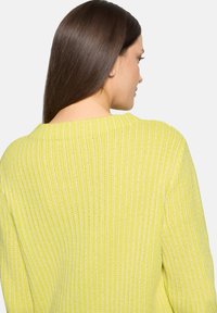 Frau mit langen dunklen Haaren, die einen leuchtend gelben, gerippten Strickpullover trägt, von hinten gezeigt und leicht zur Seite gedreht.