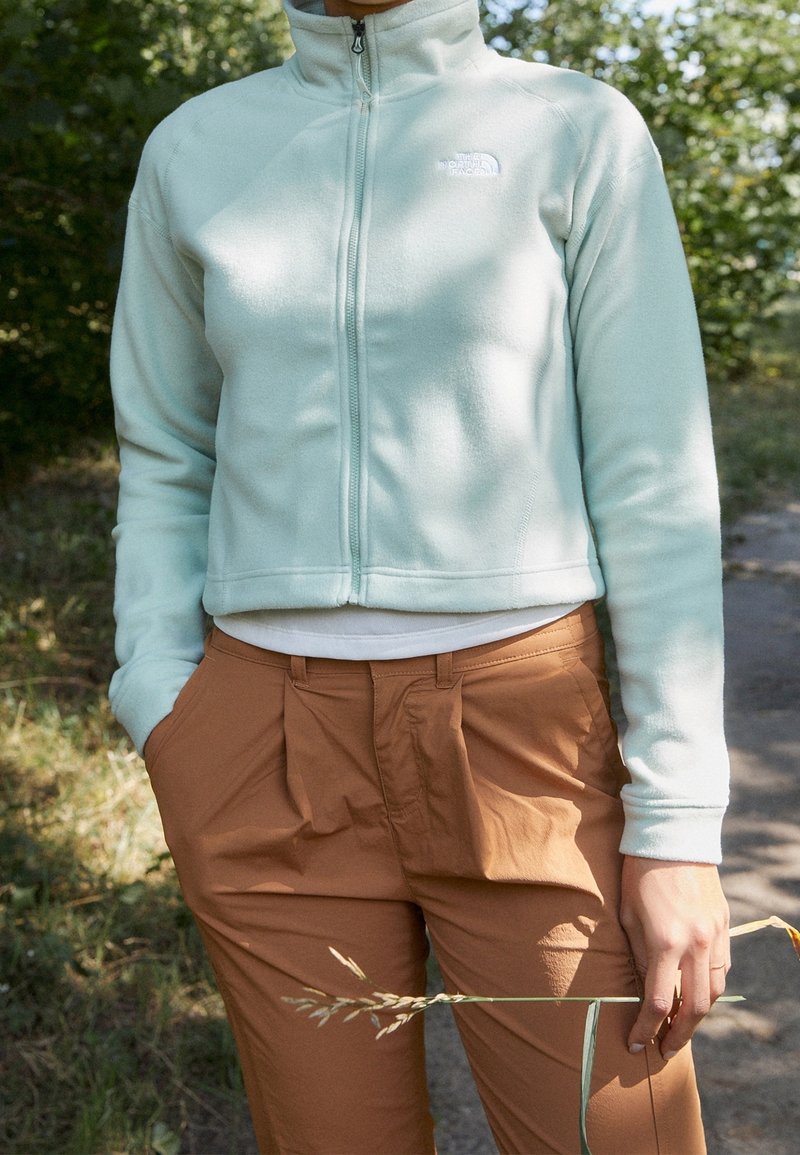 Giacca in pile verde menta chiaro con zip frontale, colletto alto e logo. Abbinata a pantaloni marroni con pieghe. Lo sfondo presenta della vegetazione.