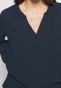 Blusa blu navy con bottoni, colletto alla cinese e due tasche sul petto, indossata da una persona con capelli castani di lunghezza media.