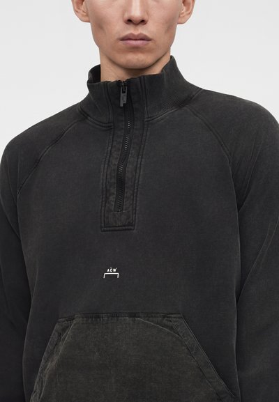 Άνδρας που φοράει σκούρο γκρι φούτερ με φερμουάρ στο στήθος, τσέπη καγκουρό και μικρό λευκό λογότυπο στο στήθος.