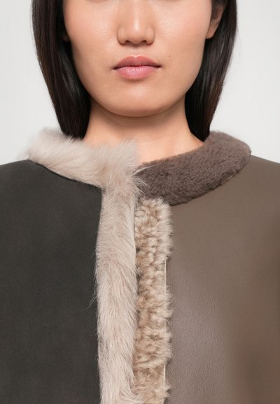 Guler din blană artificială de culoare bej deschis și maro pe un palton maro închis. Textură moale, design minimalist și detaliu de închidere asimetric.