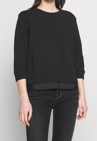 Černá mikina s tříčtvrtečními rukávy, kulatým výstřihem a texturovaným páskem s logem značky na lemu. Kombinováno s černými džínami.