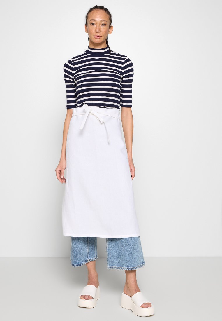 Grembiule bianco con nodo in vita, indossato sopra una maglietta a righe navy e bianche. Il modello indossa jeans blu a gamba larga cropped e scarpe bianche chunky.