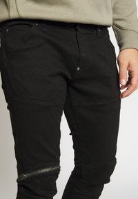 Černé kalhoty s úzkým střihem z texturované tkaniny. Mají zip na spodní části nohy a kovov é knoflíky jako ozdobu.