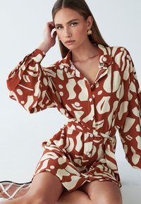 Sukienka zapinana na guziki z rdzawym tłem i kremowymi abstrakcyjnymi wzorami, długimi rękawami oraz wiązaniem w talii. Wykonana z lekkiego materiału.