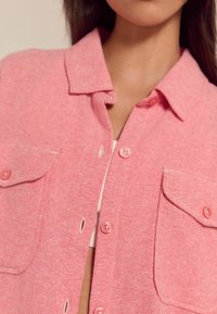 Roze katoenen shirtjack met een gestructureerd patroon, knoopsluiting en twee voorkruiszakken met bijpassende roze knopen.