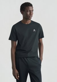 Μαύρο βαμβακερό μπλουζάκι με κοντά μανίκια, με μικρό λευκό λογότυπο Converse στο αριστερό στήθος. Απλός, κλασικός σχεδιασμός κατάλληλος για καθημερινή χρήση.